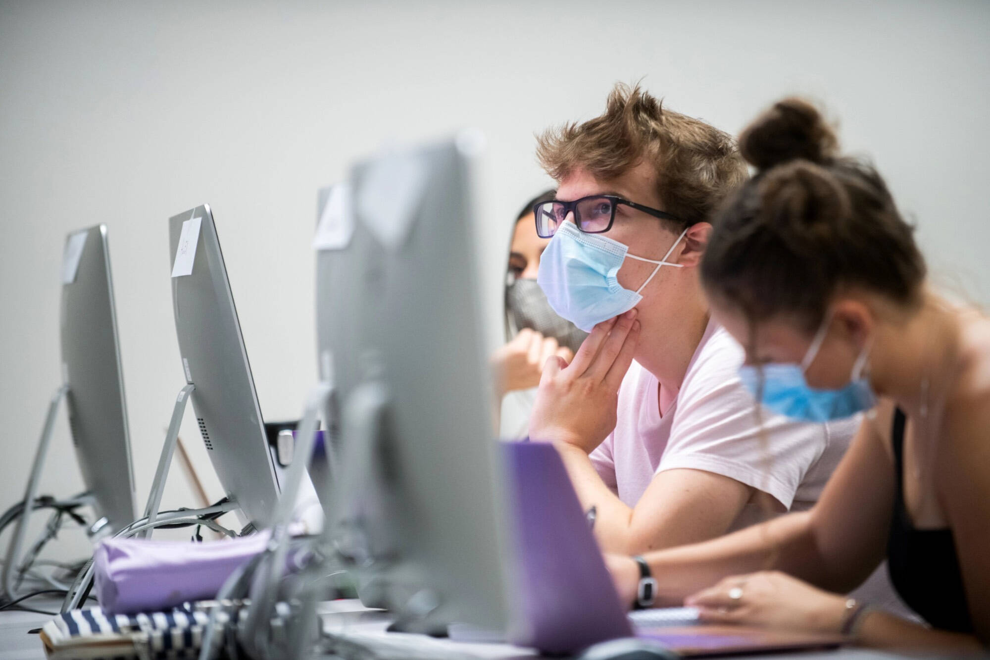 Students sit alongside a table with silver computers in front of them. One is writing and the others look at the monitor.